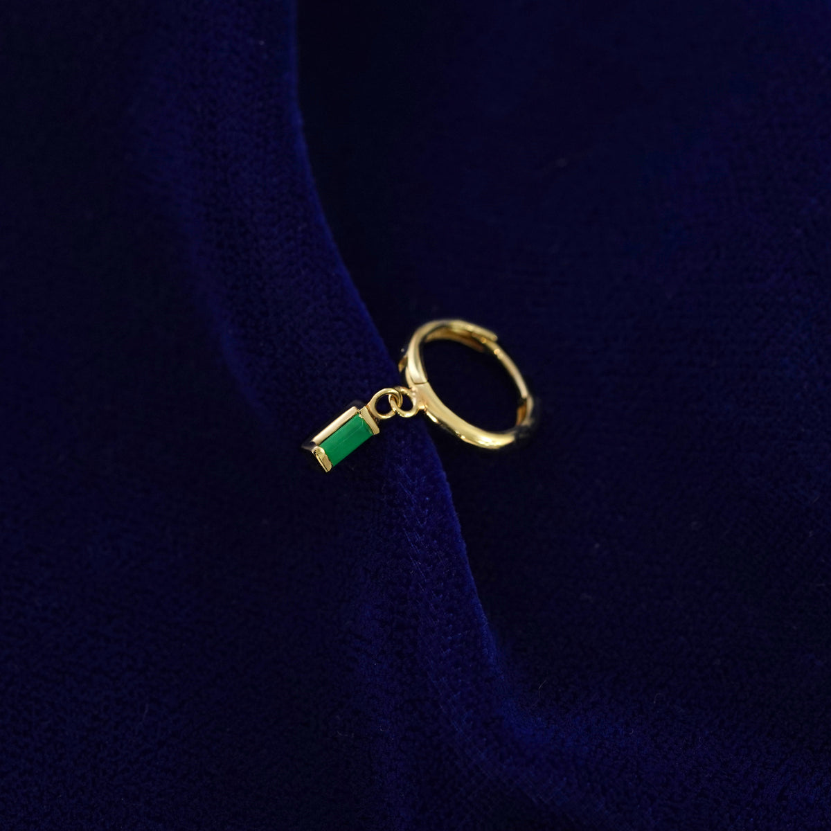 A recycled solid 14k gold Baguette Emerald Dangle Huggie Hoop earring on a dark blue background