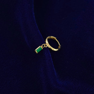 A recycled solid 14k gold Baguette Emerald Dangle Huggie Hoop earring on a dark blue background