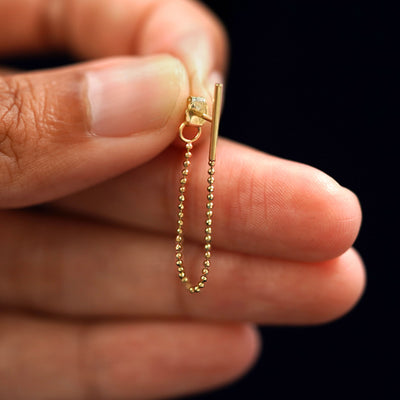 A solid yellow gold Line Dangle with the backing attached to the post held between to a model's fingers