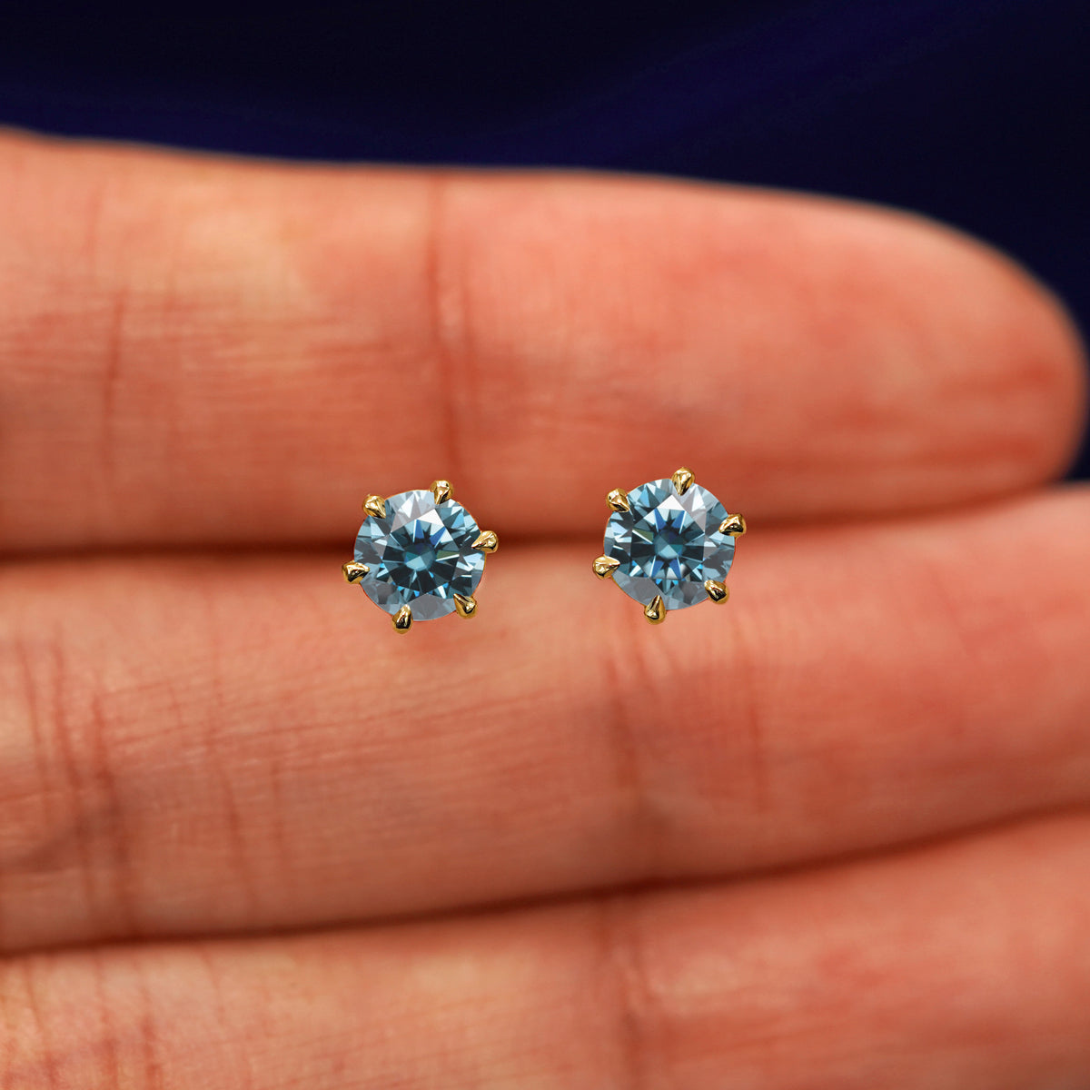A pair of solid yellow gold blue 5mm Moissanite Earrings in between a model's fingers