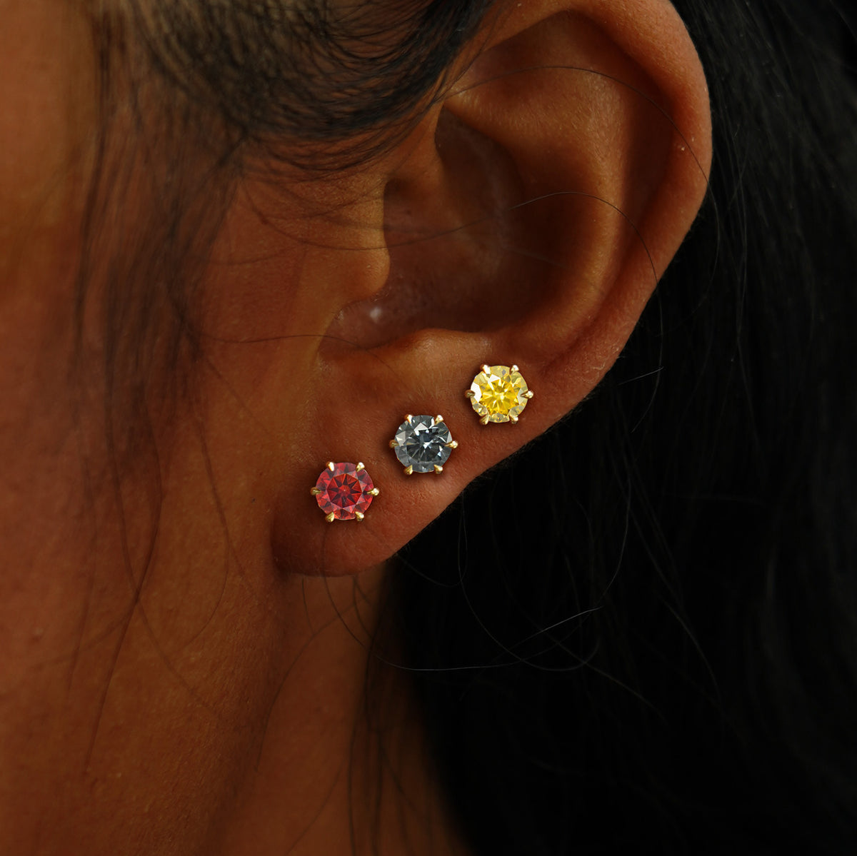 A model's ear wearing 14k gold red, grey, and yellow 5mm Moissanite stud earrings in three lobe piercings