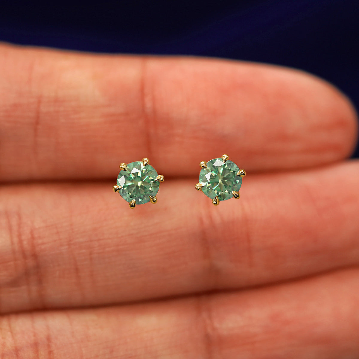 A pair of solid yellow gold green 5mm Moissanite Earrings in between a model's fingers