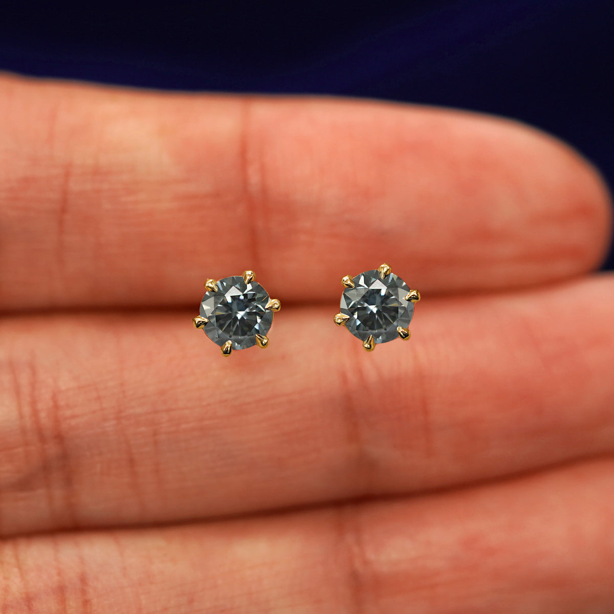 A pair of solid yellow gold grey 5mm Moissanite Earrings in between a model's fingers
