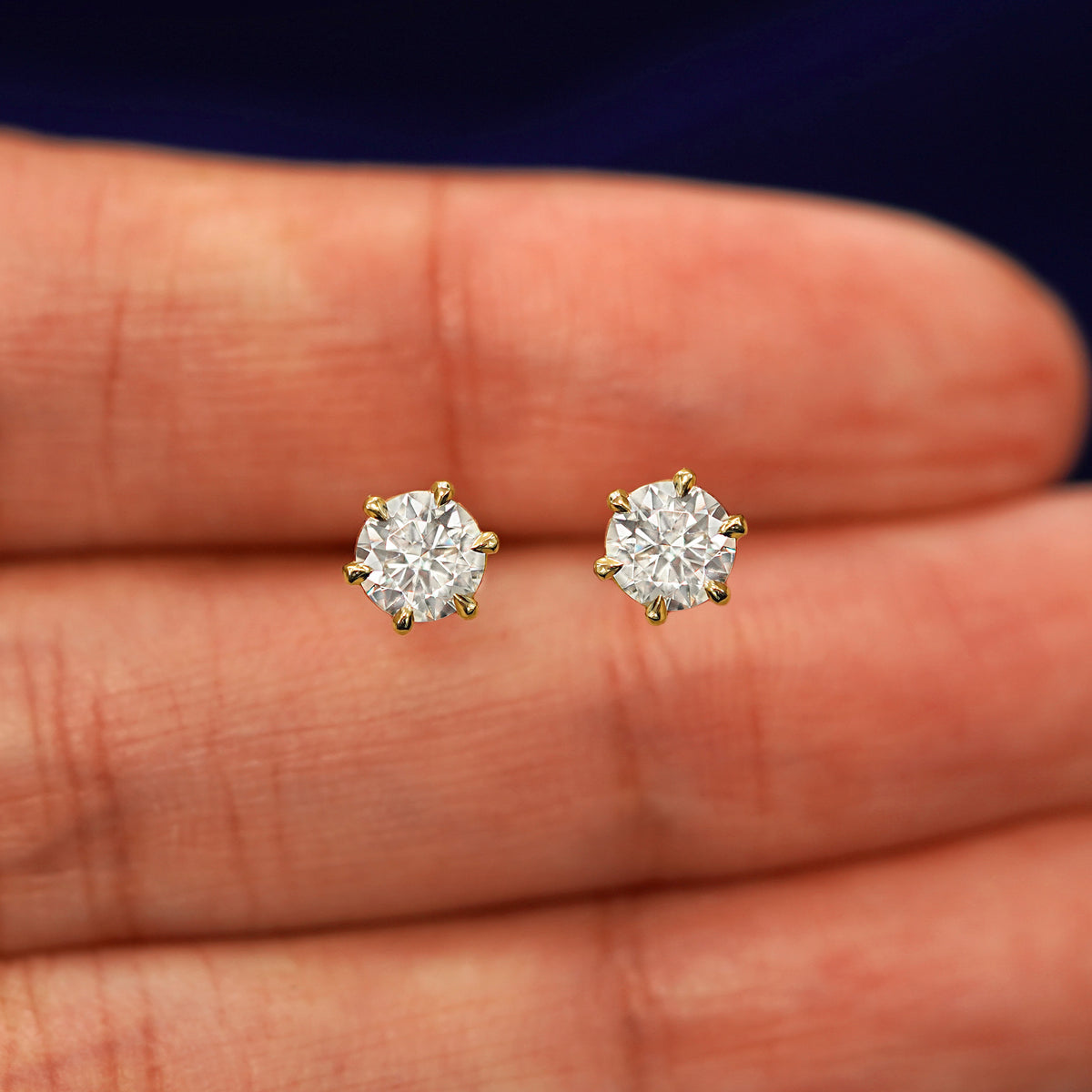 A pair of solid yellow gold clear 5mm Moissanite Earrings in between a model's fingers