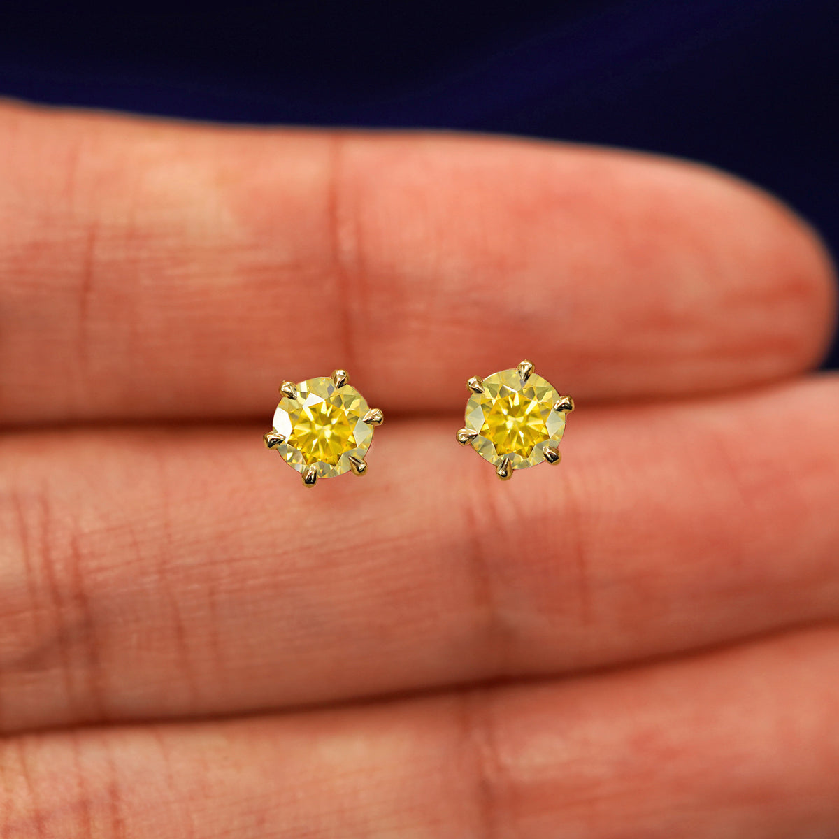 A pair of solid yellow gold yellow 5mm Moissanite Earrings in between a model's fingers