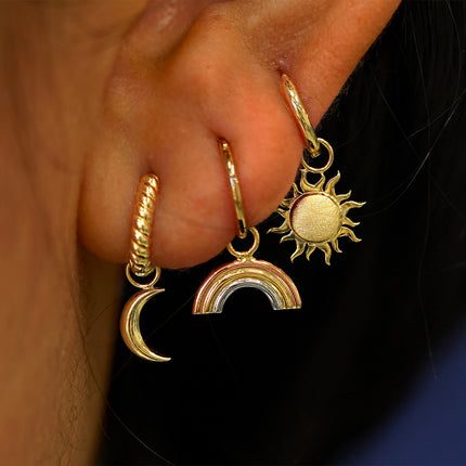 A models ear with a moon charm on a rope huggie, a rainbow charm on a seamless huggie, and a sun charm on a curvy huggie