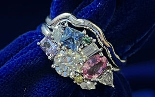 A custom cluster engagement ring made with gems of various shapes and colors and a stackable wedding band on a blue cloth