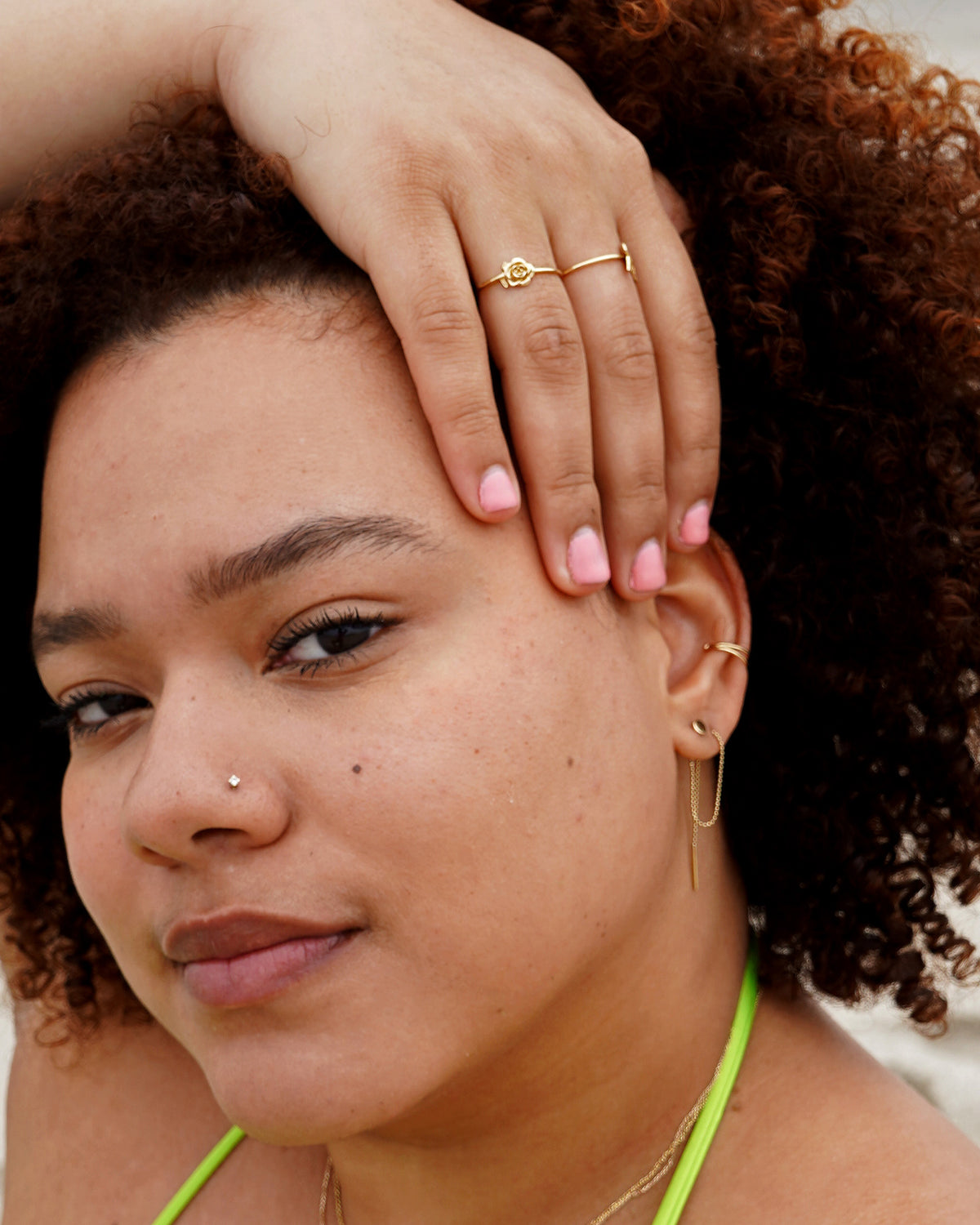 A model with their hand resting in their hair is wearing solid 14k yellow gold rings, earrings, chains and a nose stud