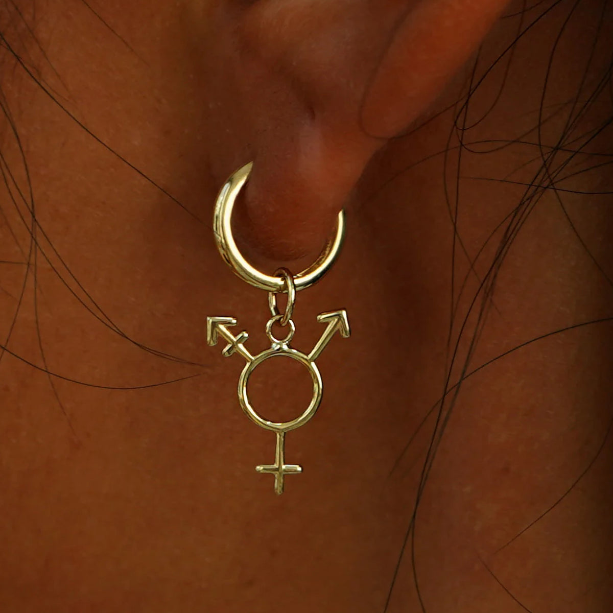 Close up view of a model's ear wearing a Small Curvy Huggie Hoop with a Trans Charm all in yellow gold