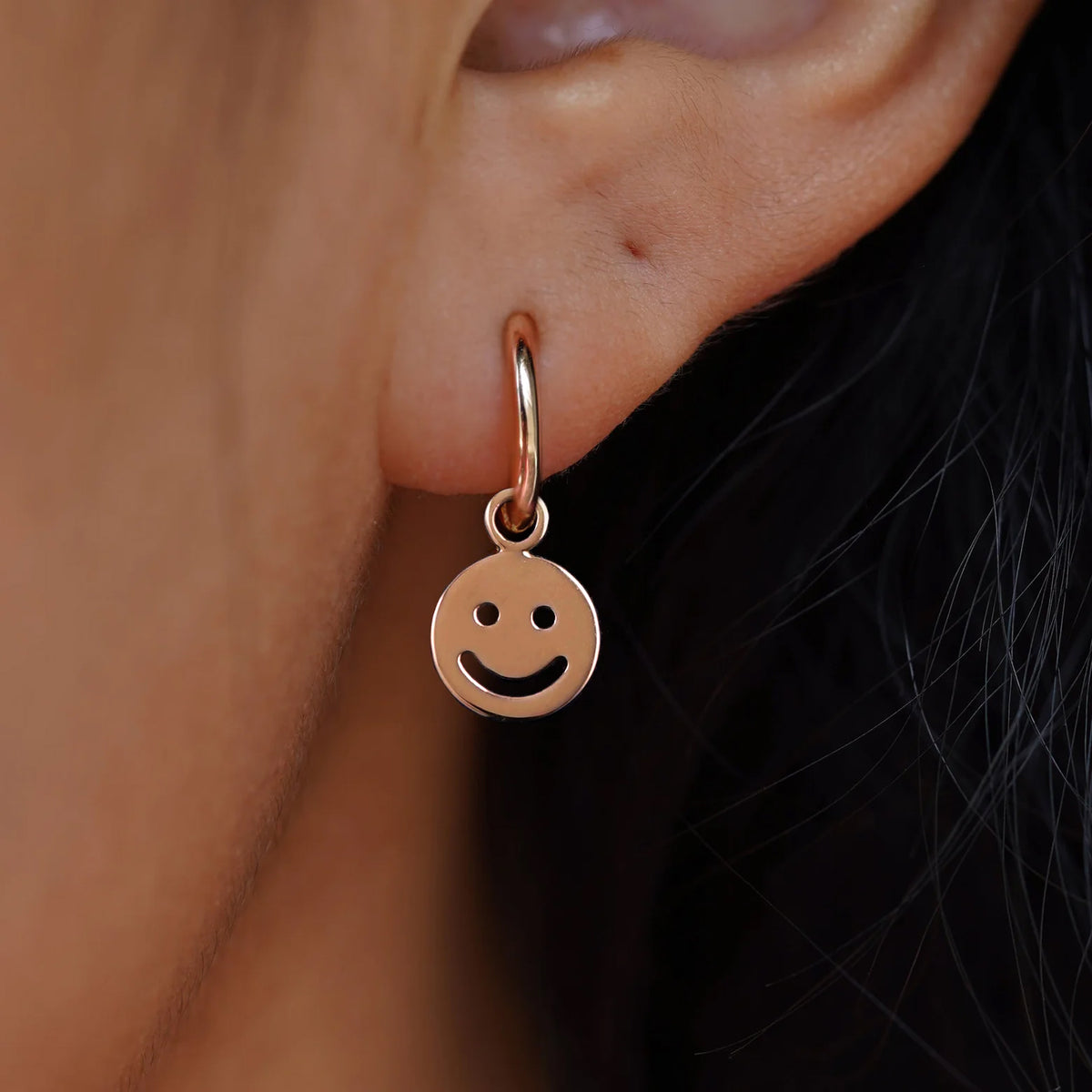 Close up view of a model's ear wearing a rose gold Smiley Face Charm on a Small Curvy Huggie Hoop