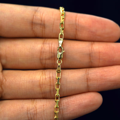 A model's hand holding a yellow 14k gold Anchor Anklet with a lobster claw clasp