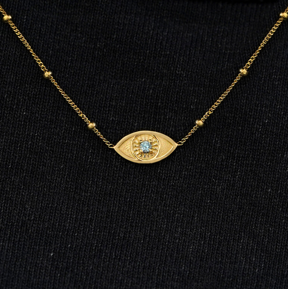 Close up view of a solid 14k yellow gold aquamarine evil eye necklace against a model's black shirt