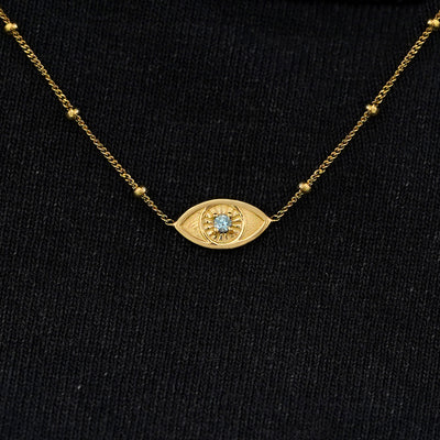 Close up view of a solid 14k yellow gold aquamarine evil eye necklace against a model's black shirt