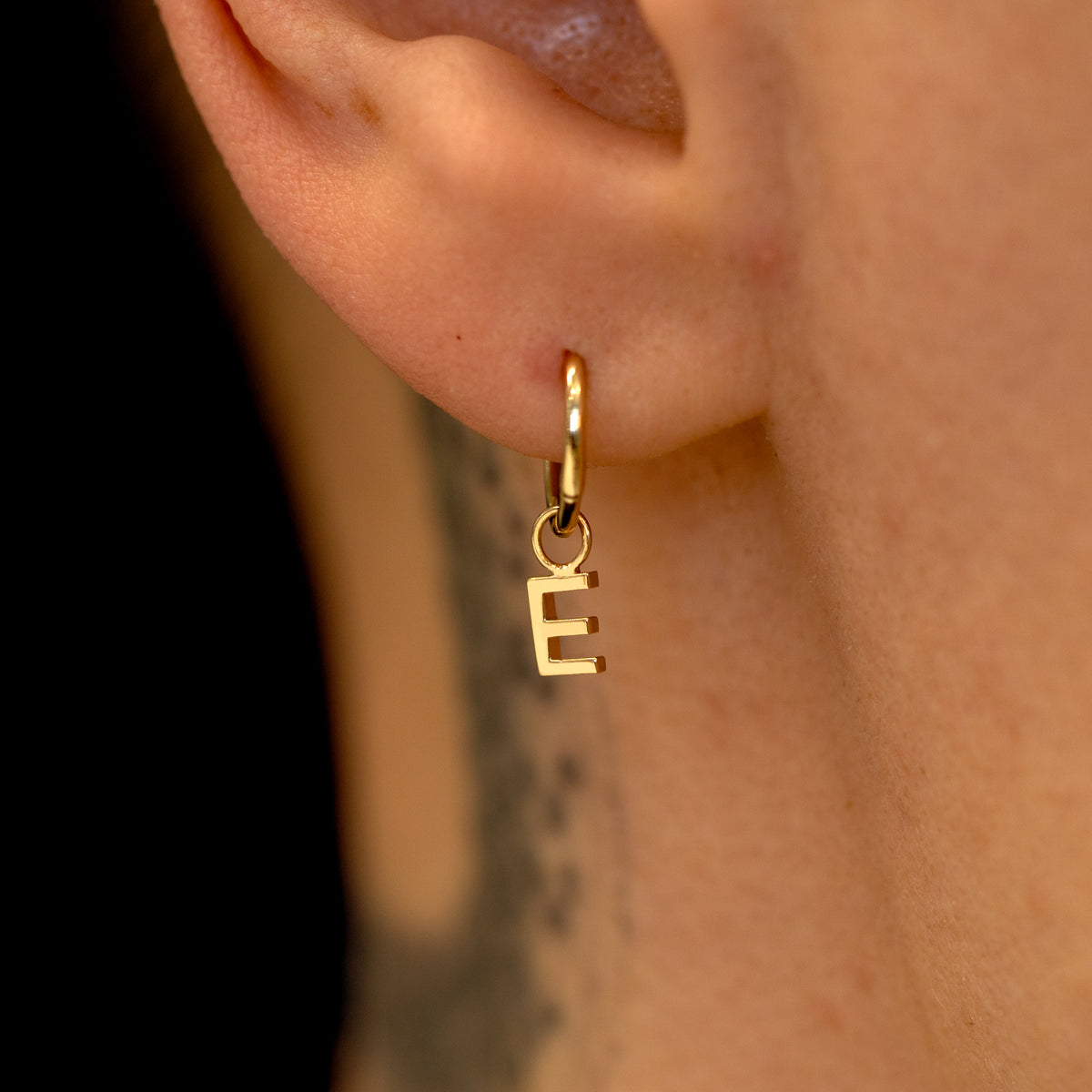 Close up view of a model's ear wearing a 14k yellow gold Seamless Huggie Hoop with a letter E block initial charm