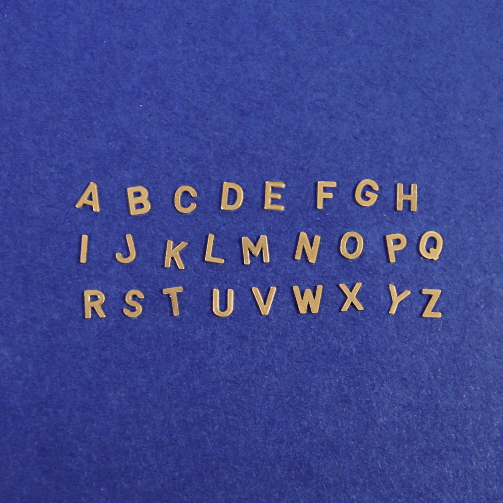 All twenty six block letters in solid 14k yellow gold laying on a dark blue background