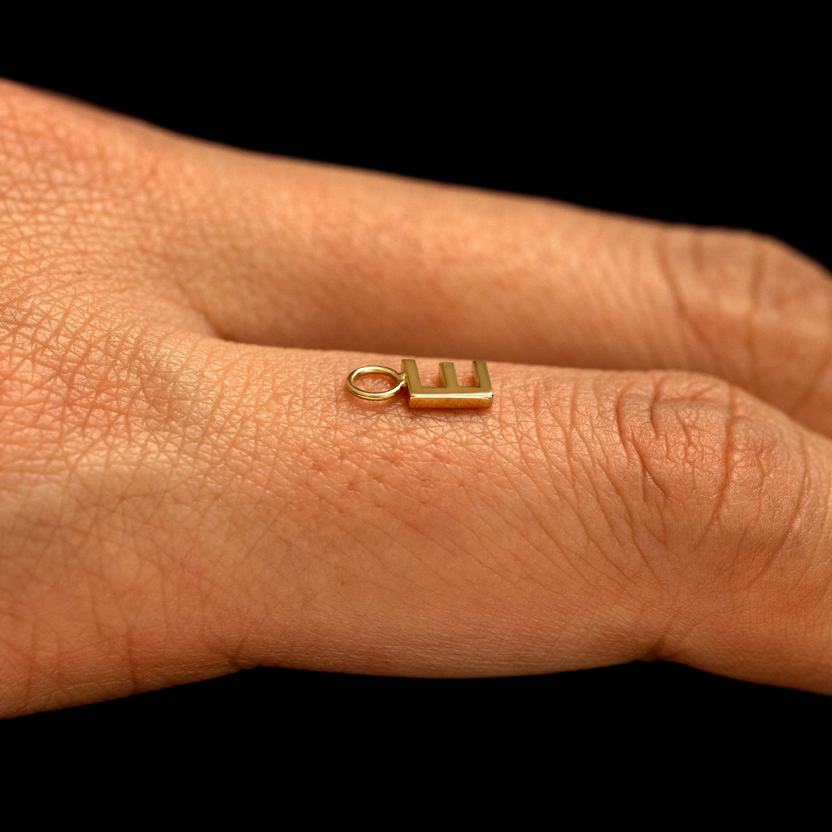 A solid 14k gold Block Initial Charm for earring in the letter E laying on the back of a model's finger