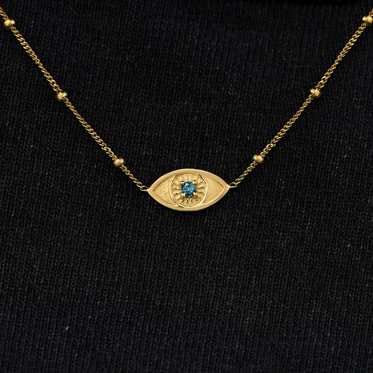 Close up view of a solid 14k yellow gold blue topaz evil eye necklace against a model's black shirt