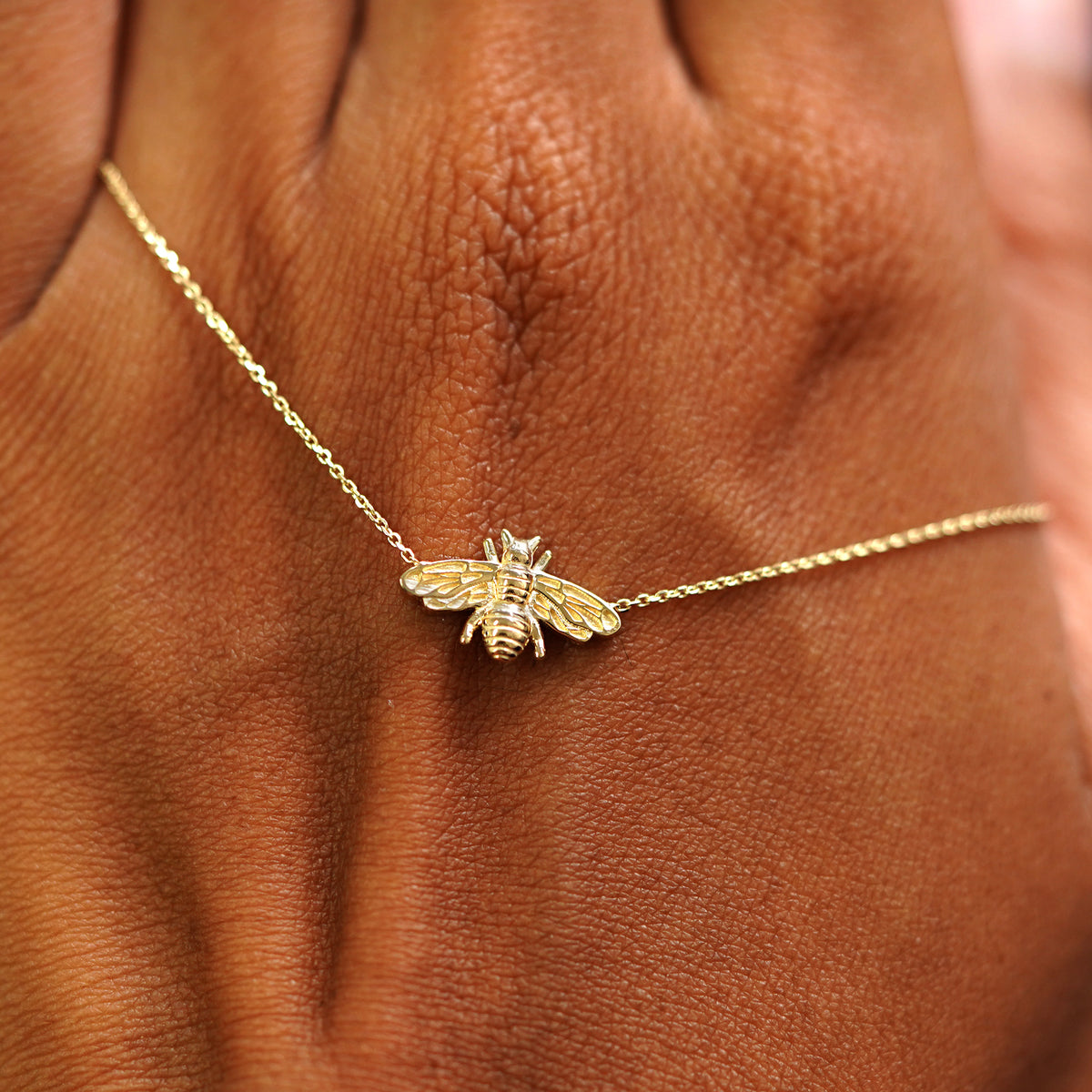 A solid gold Bee Necklace resting on the back of a model's hand