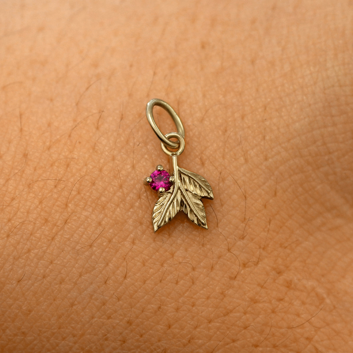 A solid 14k champagne gold ruby Gemstone 3 Leaves Charm resting on the back of a model's hand