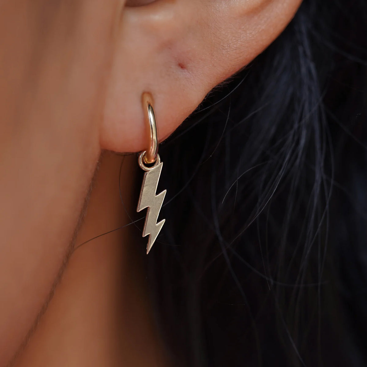 Close up view of a model's ear wearing a champagne gold Lightning Bolt Charm on a Curvy Huggie Hoop