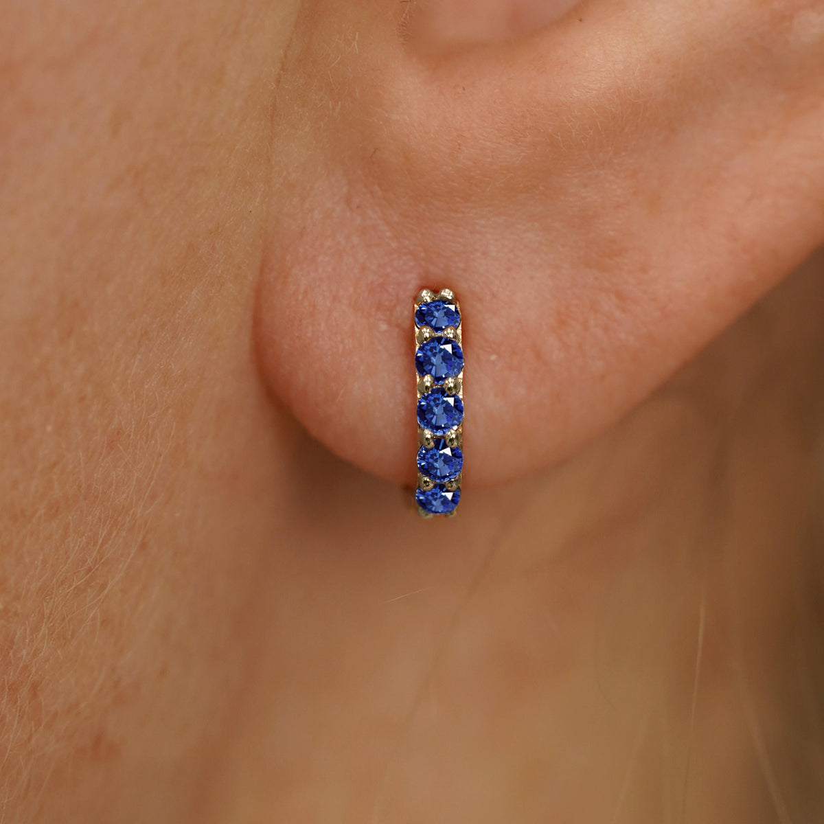 Close up view of a model's ear wearing a champagne gold Mini Blue Sapphire Huggie Hoop