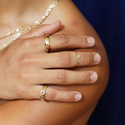 A model with their hand on their bare shoulder wearing a Curb Ring, a Tanlah Ring, a wave Ring, and a Curvy Mirror Band