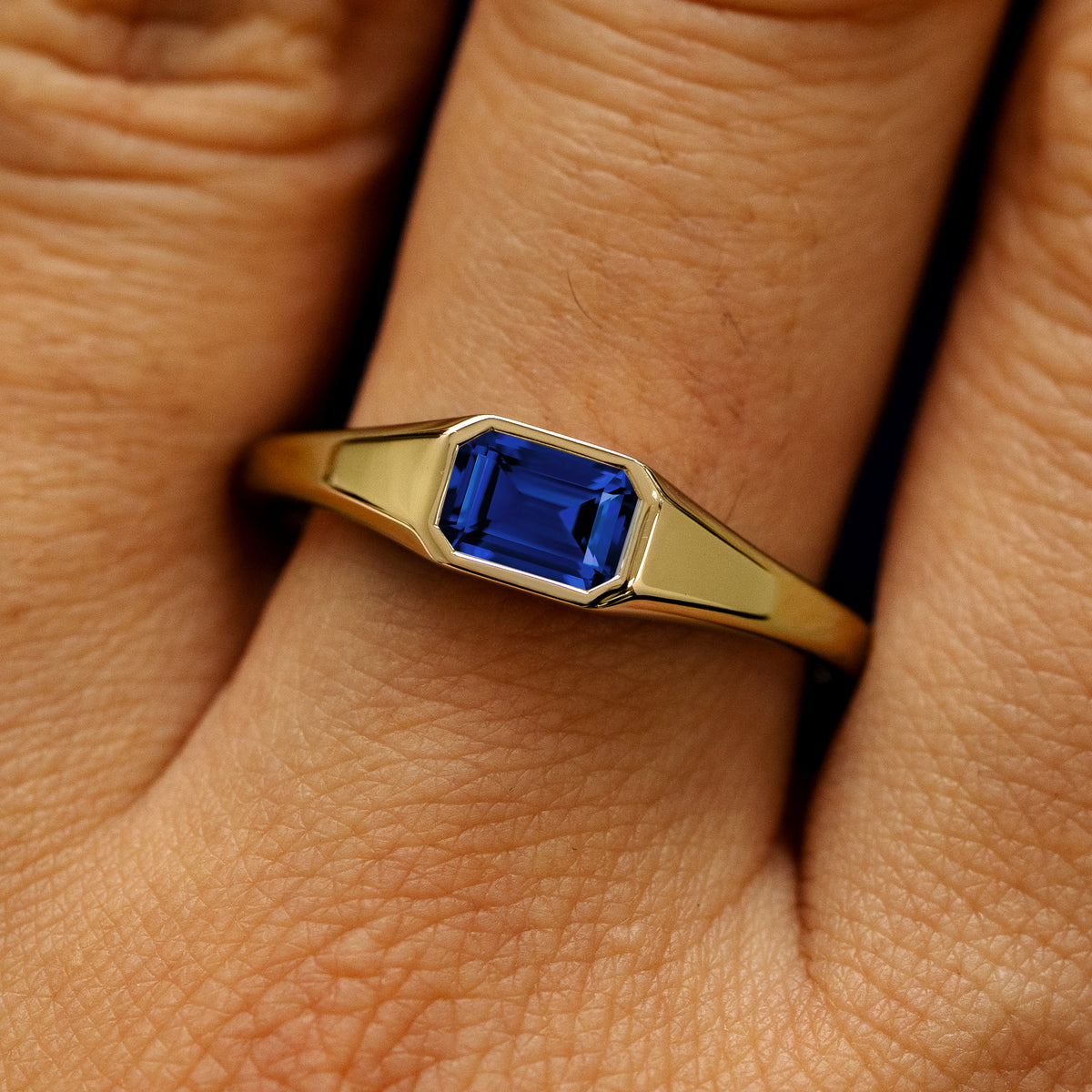 Close up view of a model's fingers wearing a 14k yellow gold Emerald Cut Lab Created Sapphire Signet Ring