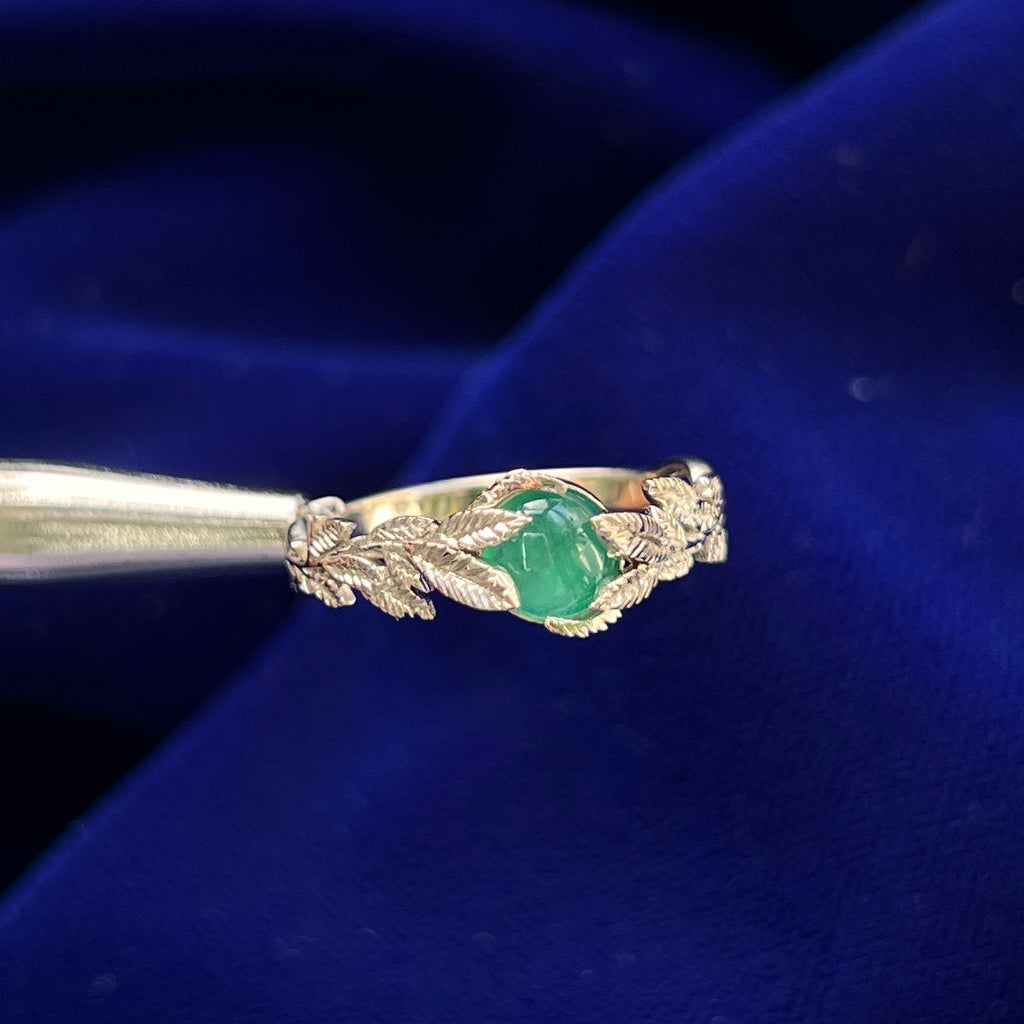 A 14 Karat yellow gold ring with double leaves hugging a cabochon oval milky emerald. The leaves are textured to resemble real leaves and look like they are spreading towards the center stone. 