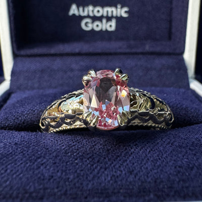 A photo of 14k Champagne Gold Double-claw-prongs-set 8x6mm Oval Peach-pink Lab Sapphire on Filigree Floral Milgrain Band Custom Ring in a blue ring box
