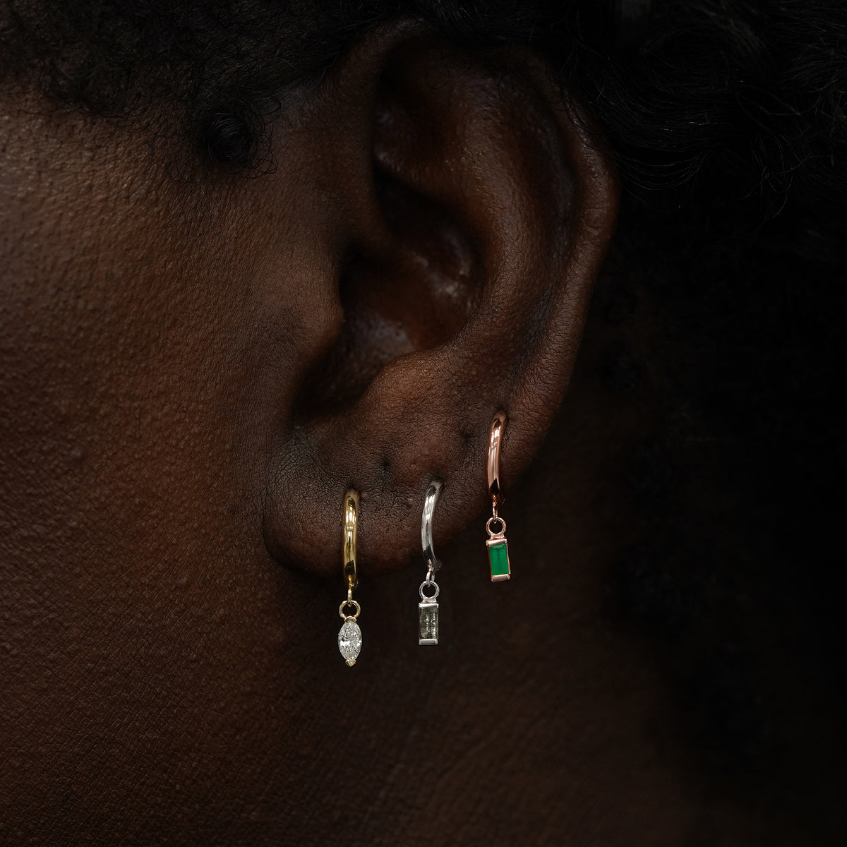 A model wearing a yellow gold marquise lab diamond, white gold baguette diamond, and rose gold baguette emerald dangle hoops