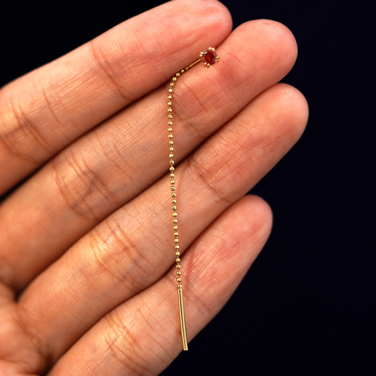 A solid gold garnet Gemstone Ear Threader Earring standing upright between a model's fingers to show the chain detail