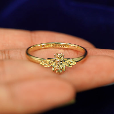 A yellow gold Curvy Bee Ring in a model's hand showing the inside of the band engraved with the words Beautifully Loved