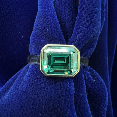 An emerald cut green moissanite solitaire ring on a dark blue cloth