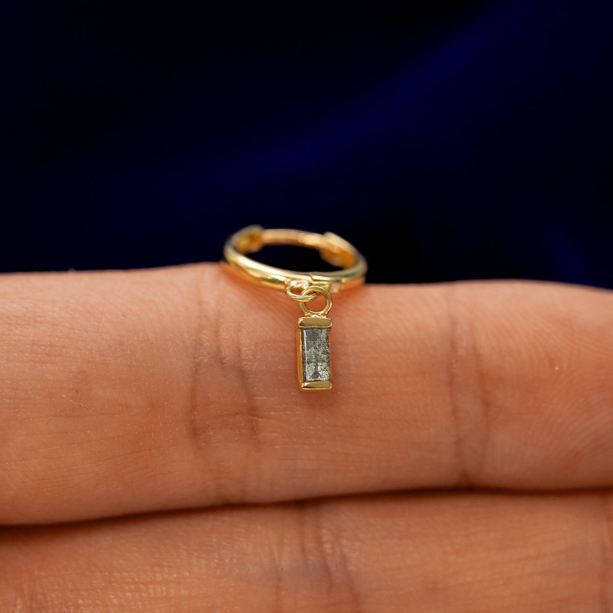 A small curvy huggie hoop earring with a dangling baguette salt and pepper charm balanced on a models finger