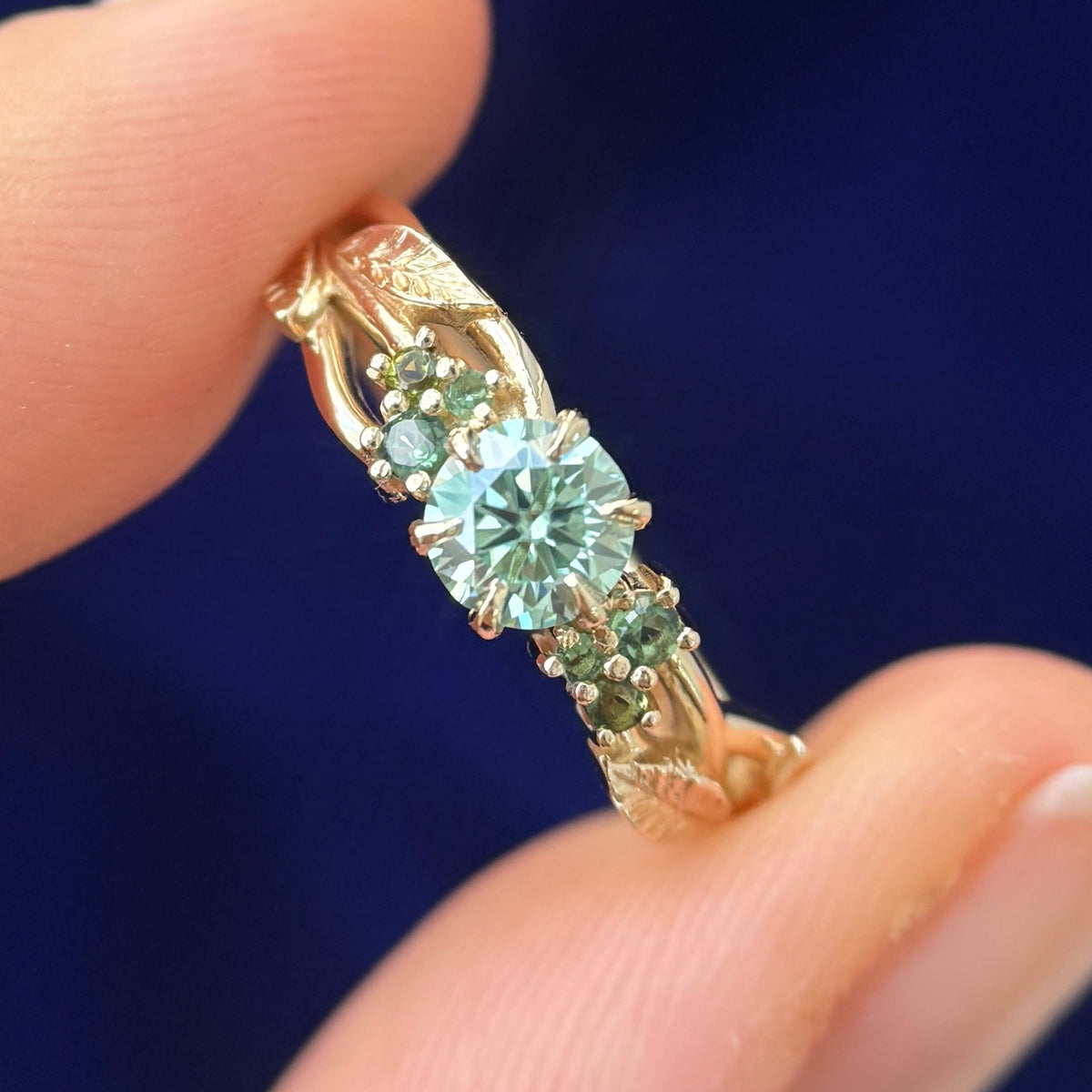 A model holding 14k Yellow Gold and 6-claw-prongs-set 5mm Round Light Green Moissanite with Green Sapphires Cluster on Leaves and Branches Custom Ring