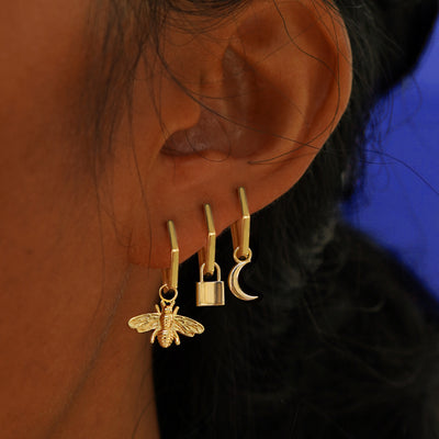 A models ear wearing three solid yellow gold Hexagon Huggie Hoop with Bee, Lock, and Moon charms in three different piercings