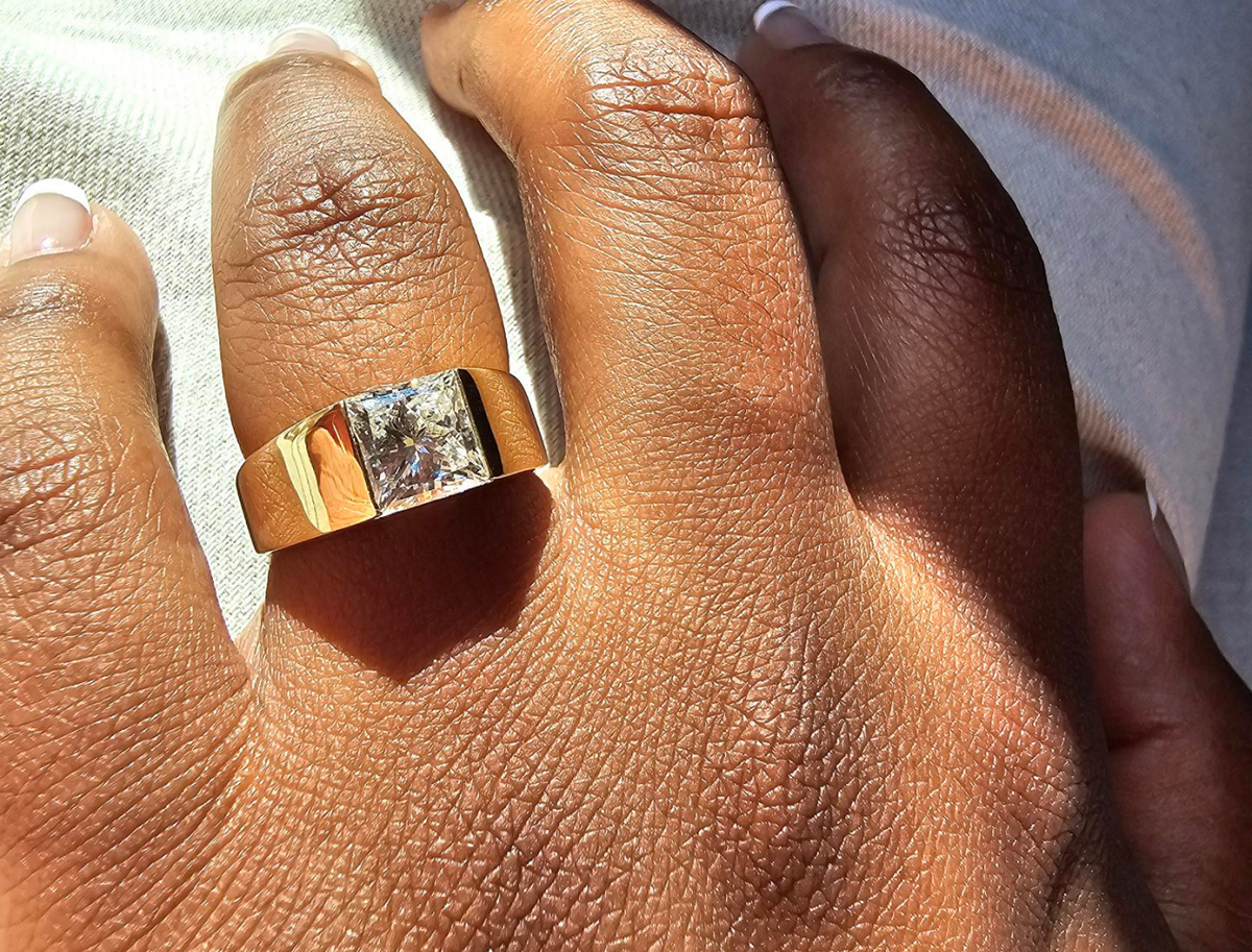 A customers hand wearing a custom engagement ring with a wide band and a princess cut diamond
