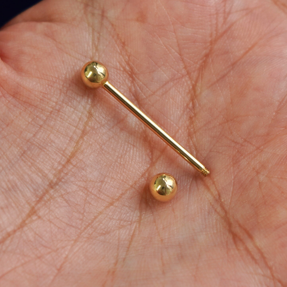 A solid yellow gold Large Straight Barbell Piercing with the externally threaded ball closure unscrewed in a model's palm