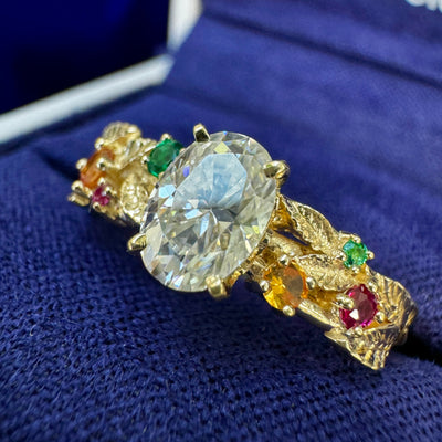 14k Yellow Gold Flower-prongs-set 7x5mm Oval Moissanite with 2 pieces Orange Sapphires, 2pc Emeralds, and 2pc Rubies on Leaves Band Custom Ring in a blue box