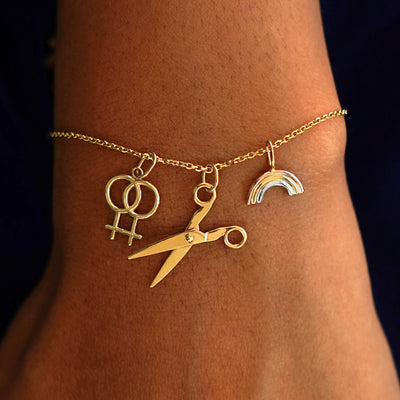 A model's wrist wearing a yellow gold Medium Cable Chain with a Lesbian Charm, a Scissors Charm, and a Rainbow Charm
