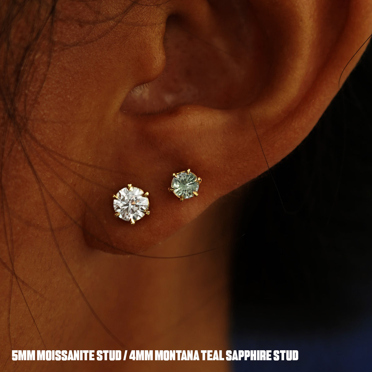 A model's ear wearing a 14k yellow gold clear 5mm Moissanite Earring and a Montana Teal Sapphire Earring in two lobe piercings