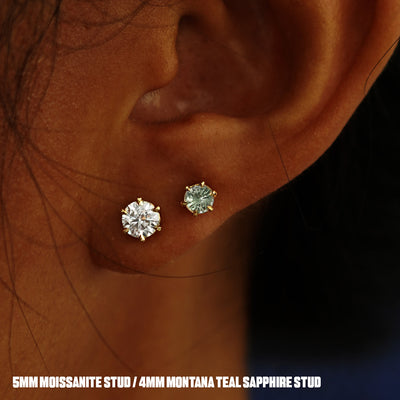 A model's ear wearing a 14k yellow gold clear 5mm Moissanite Earring and a Montana Teal Sapphire Earring in two lobe piercings