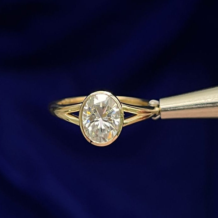 A yellow gold ring with band that features a bezel set center oval moissanite on a band with a small split on each side of the band. 