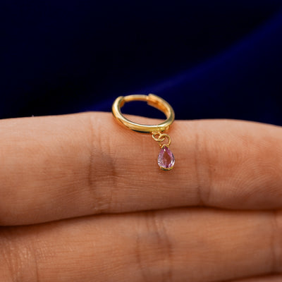 A small curvy huggie hoop earring with a dangling pear pink sapphire charm balanced on a models finger