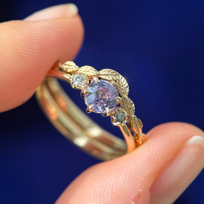 A model holding a 14k Yellow Gold and 6-claw-prongs-set 5mm Round Light Purple Sapphire with 2mm Salt-and-pepper Diamonds on 1.1-3mm Tapered Band 3-stone Custom Ring with Chevron Leaves Band