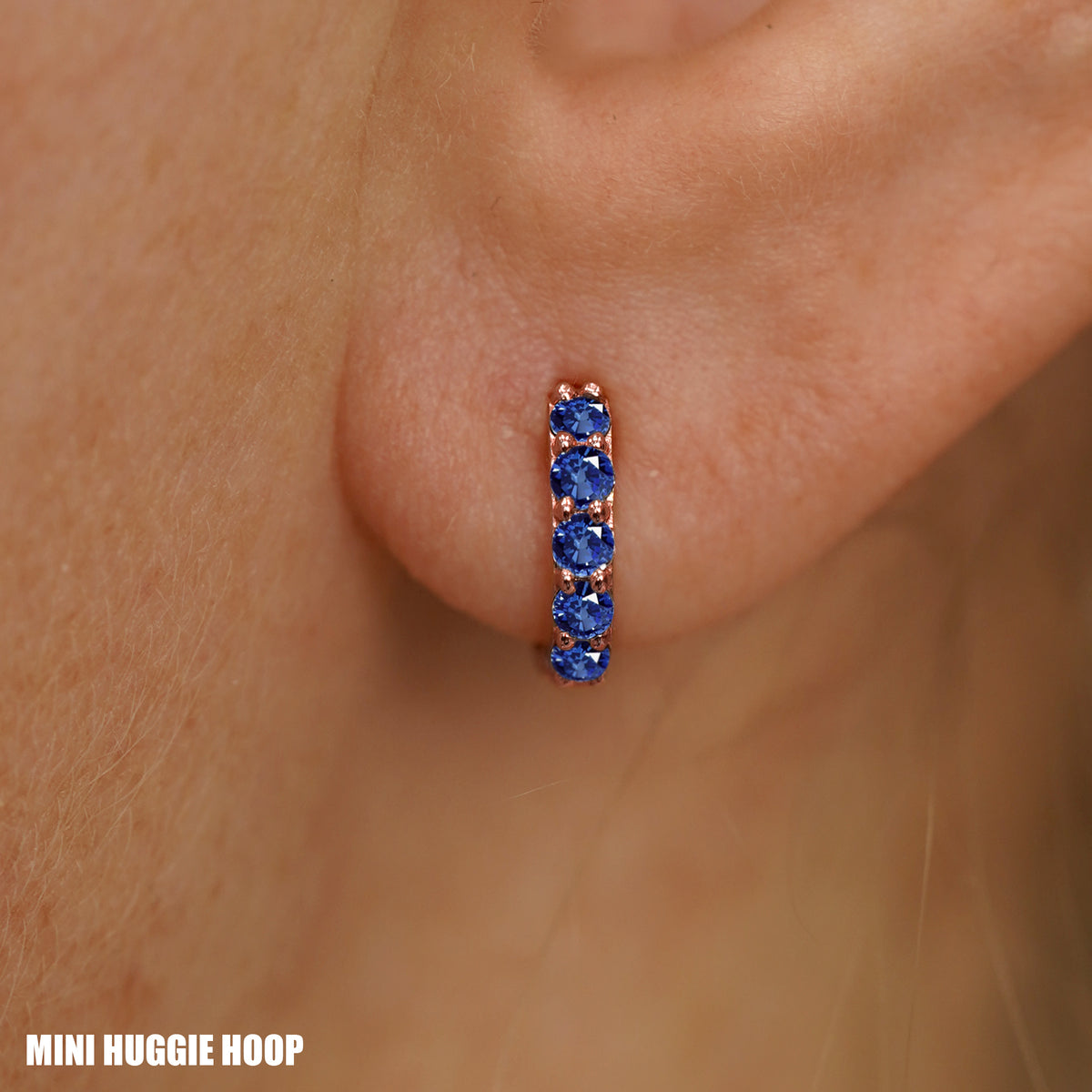 Close up view of a model's ear wearing a rose gold Mini Blue Sapphire Huggie Hoop