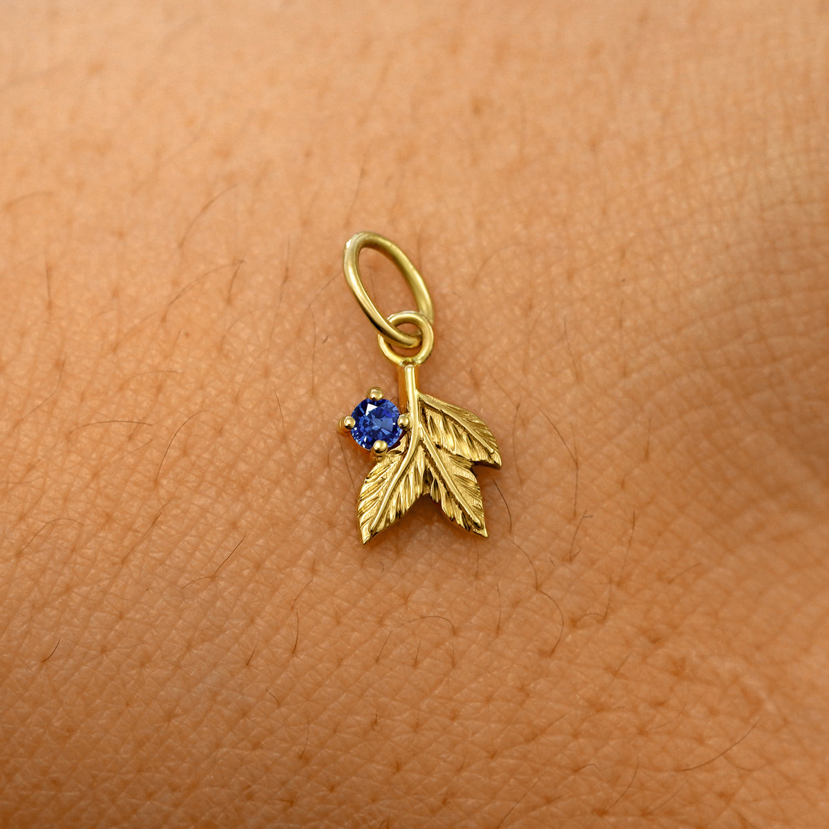 A solid 14k yellow gold blue sapphire Gemstone 3 Leaves Charm resting on the back of a model's hand