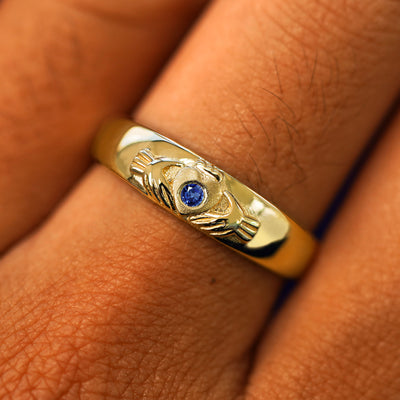 Close up view of a model's fingers wearing a 14k yellow gold blue sapphire Gemstone Claddagh Band