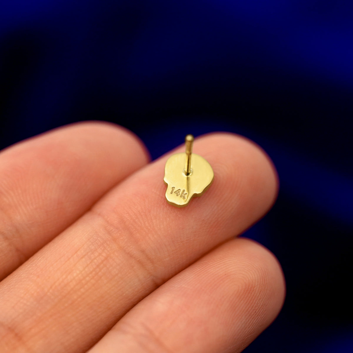 A 14k gold Skull Earring in a model's hand resting on its face  to show the back of the stud and the post