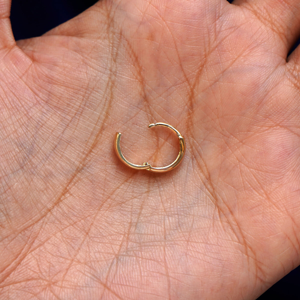A solid 14k yellow gold Small Thick Huggie Hoop Earring open in a model's palm to show the hinge